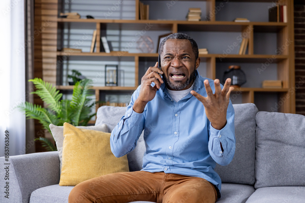 Frustrated man holding a phone while sitting on a sofa. He looks upset and is gesturing with his hand in a modern living room. Frustrated man holding a phone while sitting on a sofa. He looks upset and is gesturing with his hand in a modern living room.