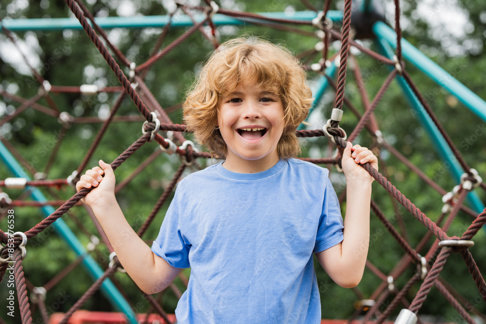 The outdoor playground for children in summer park. Kid play on playground under the tree. Portrait of cute blonde kid doing rock climbing with greenery in the background. The outdoor playground for children in summer park. Kid play on playground under the tree. Portrait of cute blonde kid doing rock climbing with greenery in the background.