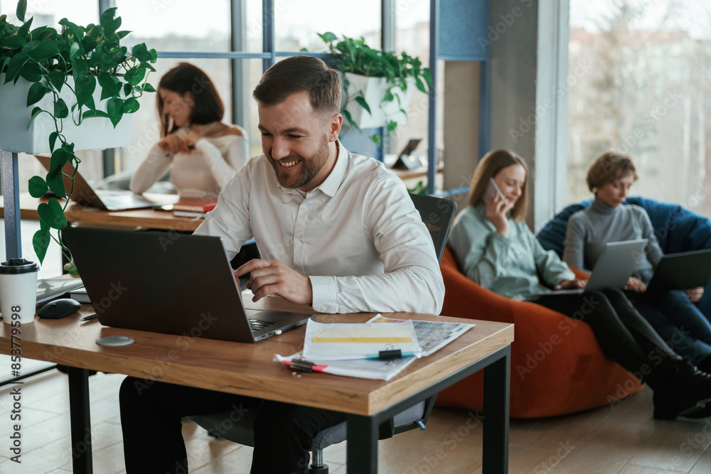 Cheerful friendly man is sitting by the table with laptop. People are working in the office with bean bags chairs in it Cheerful friendly man is sitting by the table with laptop. People are working in the office with bean bags chairs in it