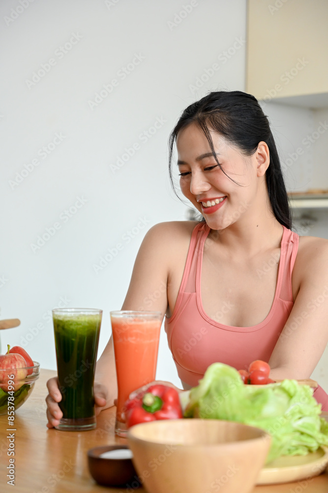 A portrait of a beautiful Asian woman in gym clothes is smiling at the camera with a glass of healthy green smoothie on a kitchen table in the kitchen, making healthy green smoothie. A portrait of a beautiful Asian woman in gym clothes is smiling at the camera with a glass of healthy green smoothie on a kitchen table in the kitchen, making healthy green smoothie.