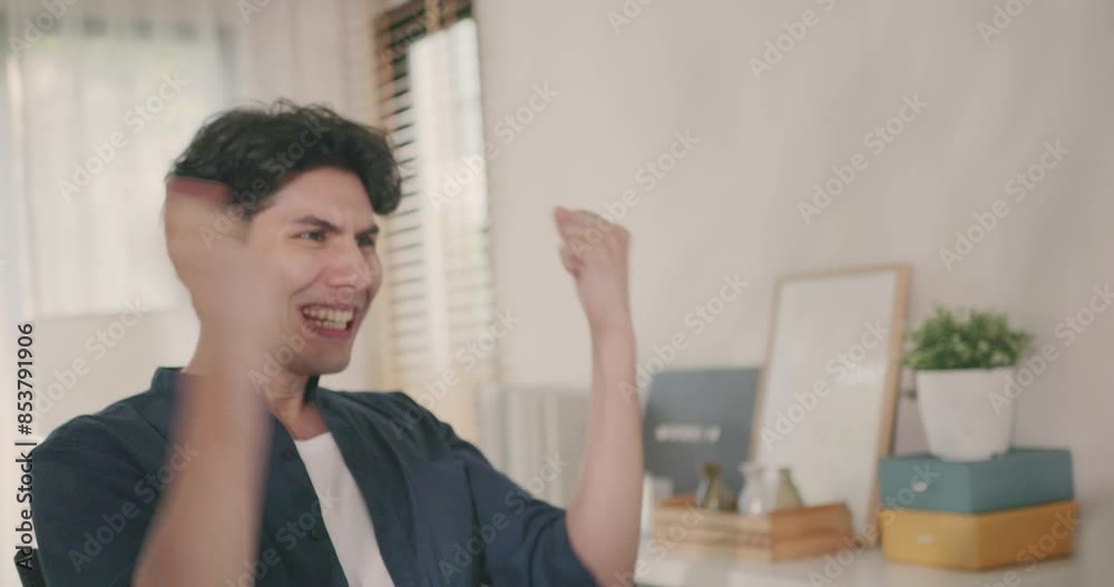 A young Asian businessman, dressed in casual attire, celebrates a major achievement with raised fists and a jubilant expression while working from his home office. A young Asian businessman, dressed in casual attire, celebrates a major achievement with raised fists and a jubilant expression while working from his home office.