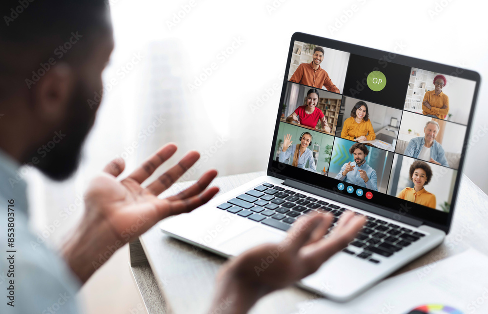 Black man sits in front of a laptop, participating in a video conference meeting with several colleagues. He has his hands raised as if he is making a point during the call. Black man sits in front of a laptop, participating in a video conference meeting with several colleagues. He has his hands raised as if he is making a point during the call.