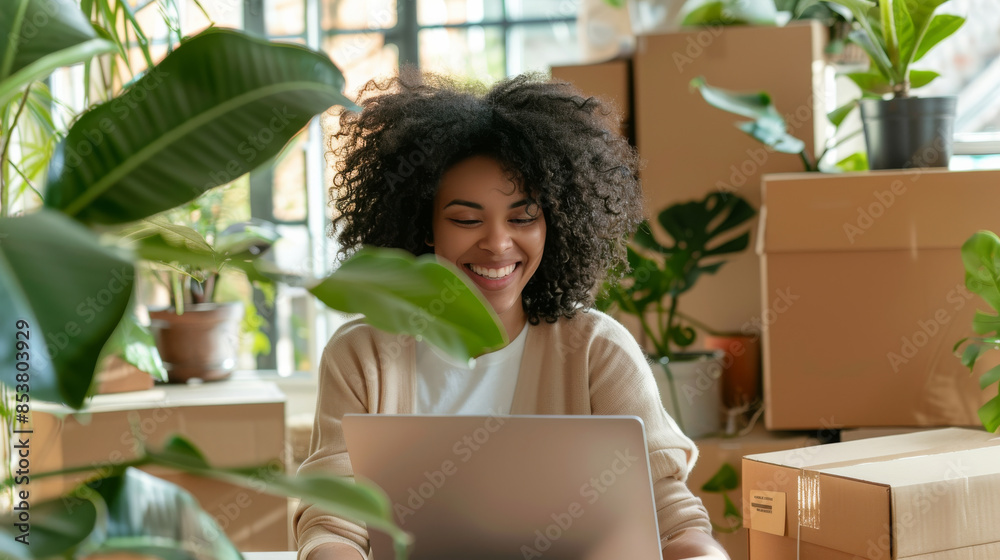 A young woman joyfully works on her laptop in a bright room filled with various potted plants and packed boxes, suggesting she is setting up a new home office. A young woman joyfully works on her laptop in a bright room filled with various potted plants and packed boxes, suggesting she is setting up a new home office.