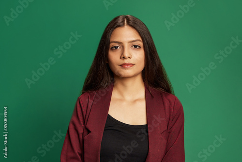 Confident young Middle-eastern woman with long dark hair wearing burgundy jacket and black top, posing against green studio background. Concept of human emotions, fashion and style, business.