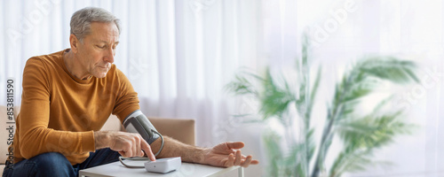 An elderly man wearing a yellow sweater sits at a table in his living room and uses a digital blood pressure monitor to check his blood pressure, copy space