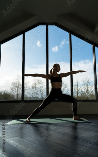 Woman is practicing ashtanga yoga vinyasa flow, warrior two asana pose exercise indoors, against the window. Wellness, lifestyle, copy space.