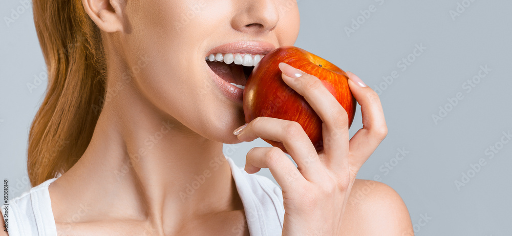 A close-up shot of a woman taking a bite out of a red apple. She is wearing a white tank top and her hair is pulled back in a ponytail. The photo is taken against a light grey background. A close-up shot of a woman taking a bite out of a red apple. She is wearing a white tank top and her hair is pulled back in a ponytail. The photo is taken against a light grey background.