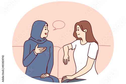 Girls from different ethnicities are talking sitting on couch demonstrating tolerance and lack of cultural prejudice. Woman in muslim clothes is talking to girlfriend of european appearance