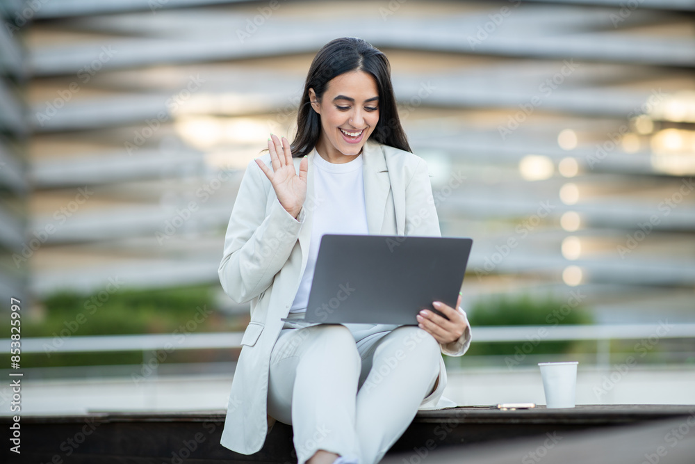 A young woman in a white suit sits on a bench outside a modern office building. She is using a laptop and waving at the camera, suggesting she is engaged in a video call A young woman in a white suit sits on a bench outside a modern office building. She is using a laptop and waving at the camera, suggesting she is engaged in a video call