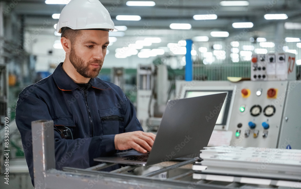 Using the laptop. Factory worker is indoors with hard hat Using the laptop. Factory worker is indoors with hard hat