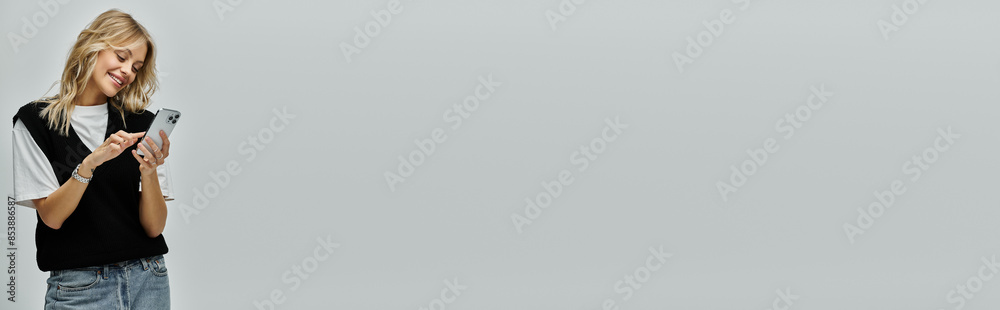 Naklejka premium A stylish young woman with blonde hair holding a cell phone in her right hand against a grey background.