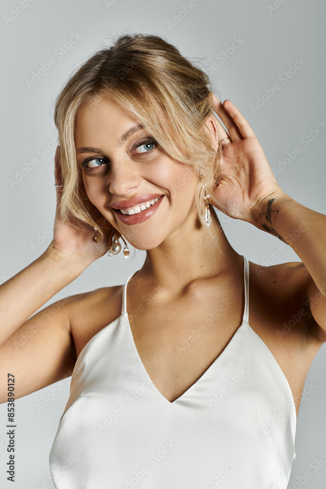A young woman with blonde hair smiles happily while wearing a white top against a grey background. A young woman with blonde hair smiles happily while wearing a white top against a grey background.