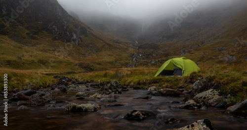 View of a green camping tent next to a river in Corrie Fee, Cairngorms National Park of Scotland, surrounded by mountains covered in mist photo