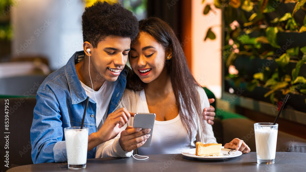 Two African American teenagers are sitting at a cozy cafe, enjoying milkshakes and a slice of cake. They are smiling and watching something on a smartphone together. Two African American teenagers are sitting at a cozy cafe, enjoying milkshakes and a slice of cake. They are smiling and watching something on a smartphone together.