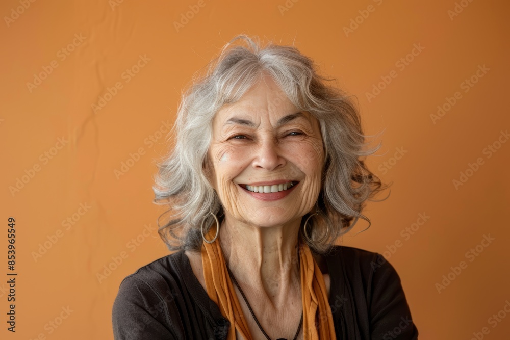 Portrait of a smiling woman in her 70s smiling at the camera while standing against pastel orange background Portrait of a smiling woman in her 70s smiling at the camera while standing against pastel orange background