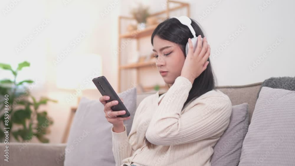 A young Asian woman is sitting on a couch, wearing headphones and holding a smartphone. The background features a well-lit living room with plants and shelves A young Asian woman is sitting on a couch, wearing headphones and holding a smartphone. The background features a well-lit living room with plants and shelves