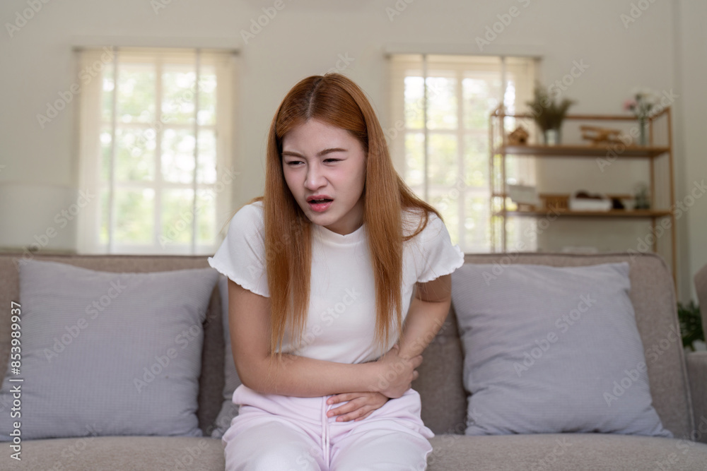 Young Woman Experiencing Stomach Pain While Sitting on a Couch in a Bright Living Room Young Woman Experiencing Stomach Pain While Sitting on a Couch in a Bright Living Room
