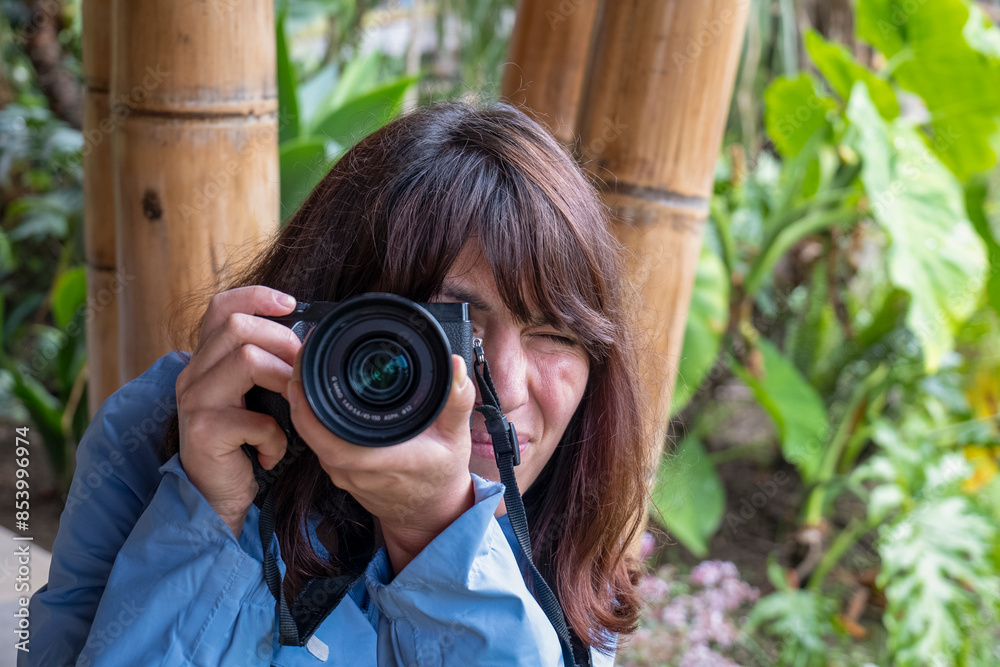 A Young Brown Woman in a Blue Coat with a Camera with Strap Around her Neck Takes a Photo Doing a Grimace on A Rainy Day with Bamboo Poles in the Background at a Telephoto Angle