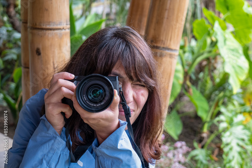Obraz na płótnie A Young Brown Woman in a Blue Coat with a Camera with Strap Around her Neck Takes a Photo Doing a Grimace on A Rainy Day with Bamboo Poles in the Background at a Telephoto Angle w salonie