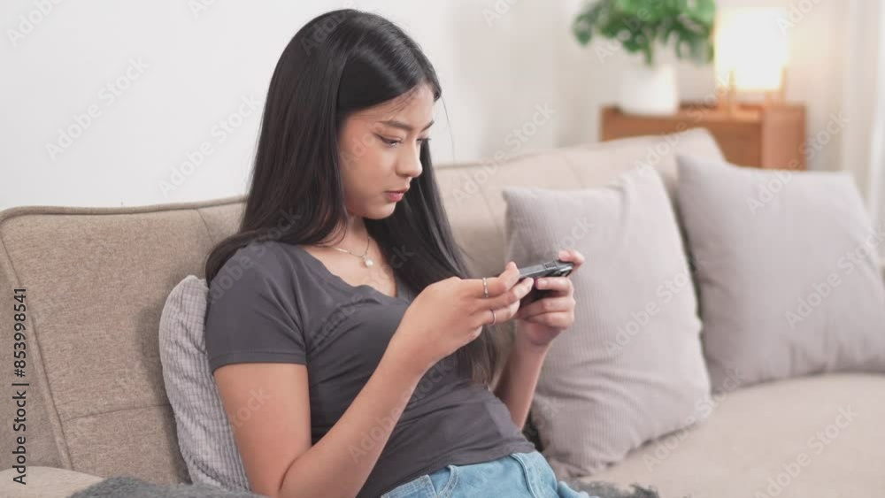 A young Asian woman is sitting on a couch, playing a game on her smartphone with an excited expression. She raises her fist in a gesture of triumph A young Asian woman is sitting on a couch, playing a game on her smartphone with an excited expression. She raises her fist in a gesture of triumph