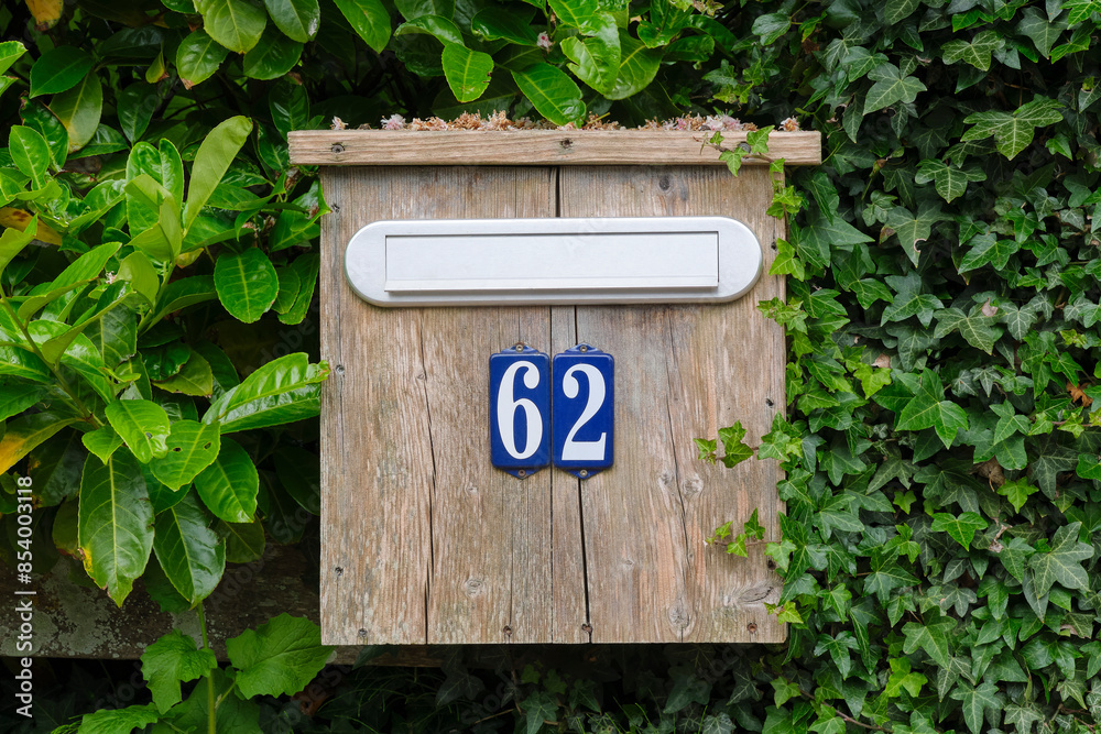 Old wooden unpainted mailbox with enamel number 62 in blue and white between green creepers Old wooden unpainted mailbox with enamel number 62 in blue and white between green creepers