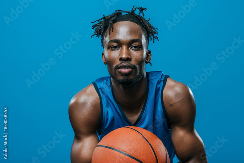 a Black man in a blue sports jersey, looking confident while holding a basketball on a blue studio background