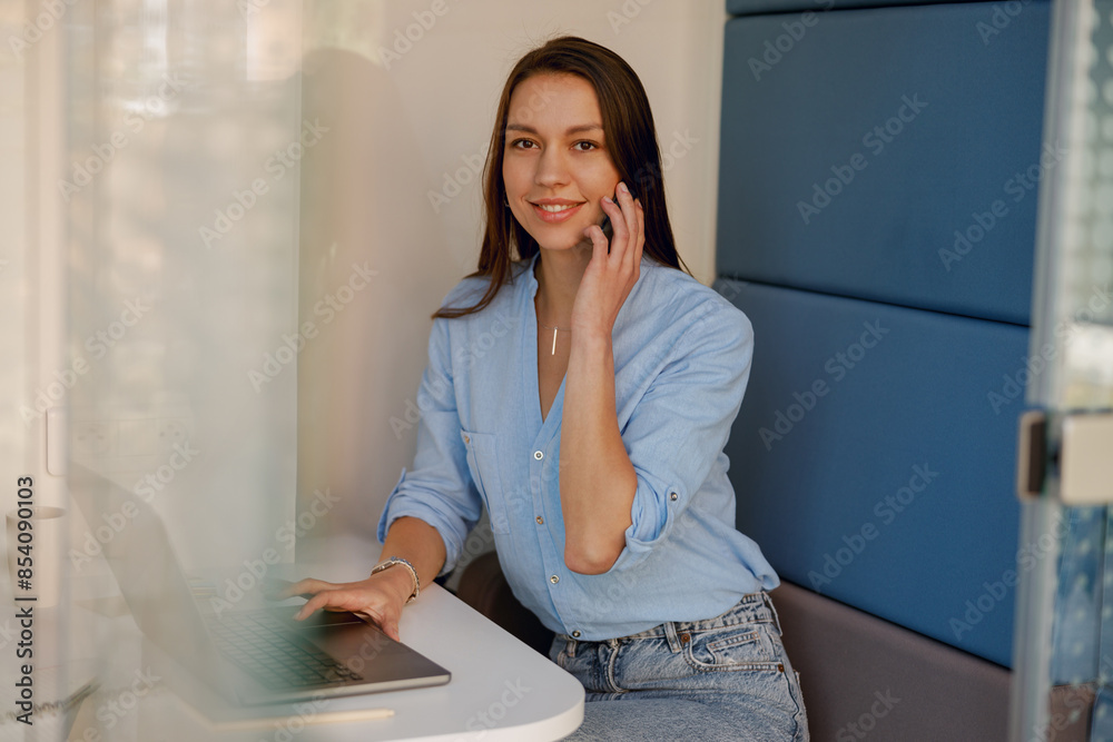 Fototapeta premium A smiling woman is having a phone call while working on a laptop in a modern office space