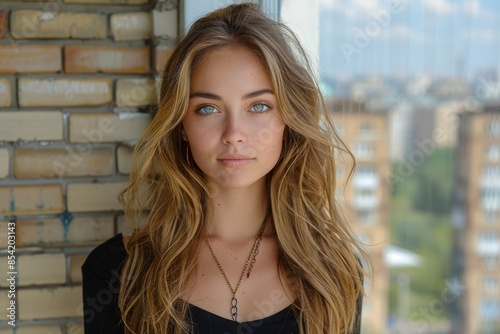 A portrait of a blue-eyed girl with long, wavy blonde hair, standing against a brick wall with a blurred cityscape in the background on a sunny day photo