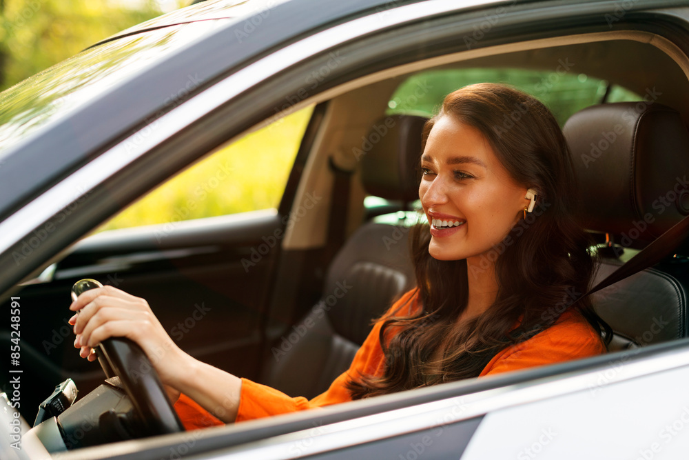 Happy smiling lady in earbuds driving car, enjoying music and drive alone, holding hands on steering wheel Happy smiling lady in earbuds driving car, enjoying music and drive alone, holding hands on steering wheel