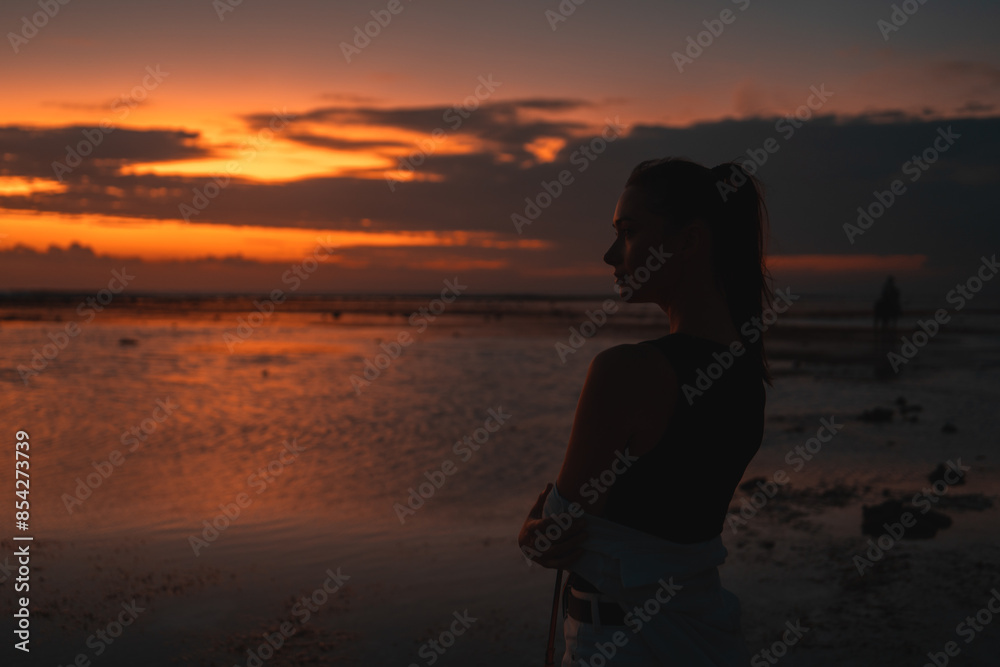 Woman contemplating the sunset by the sea, captured in silhouette. The vibrant sky and reflective water create a serene, contemplative scene, emphasizing the beauty and tranquility of the moment.