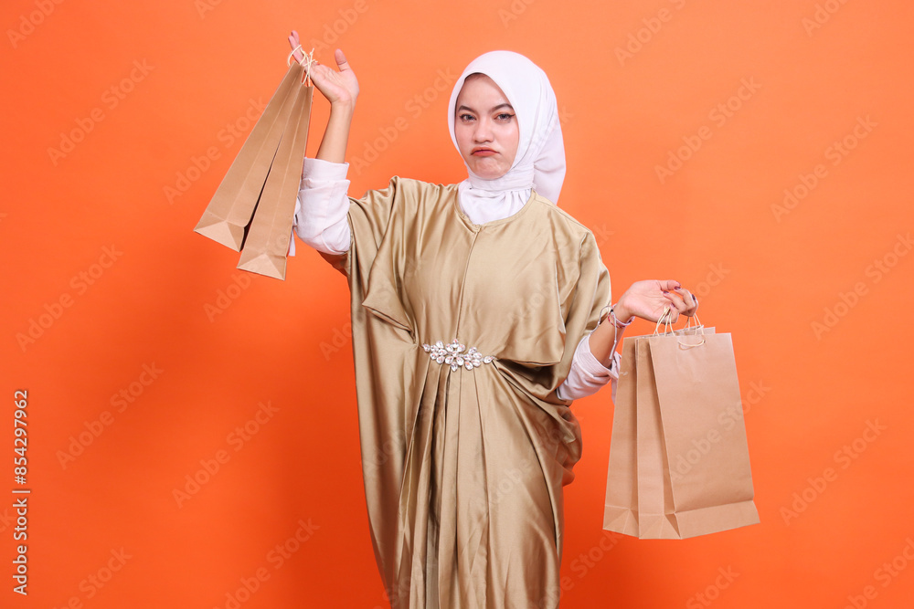 Disappointed young indonesia girl carrying and lifting aside paper bag containing groceries wearing a kaftan Muslim dress with a hijab, isolated on an orange background. concept transaction,promotion Disappointed young indonesia girl carrying and lifting aside paper bag containing groceries wearing a kaftan Muslim dress with a hijab, isolated on an orange background. concept transaction,promotion