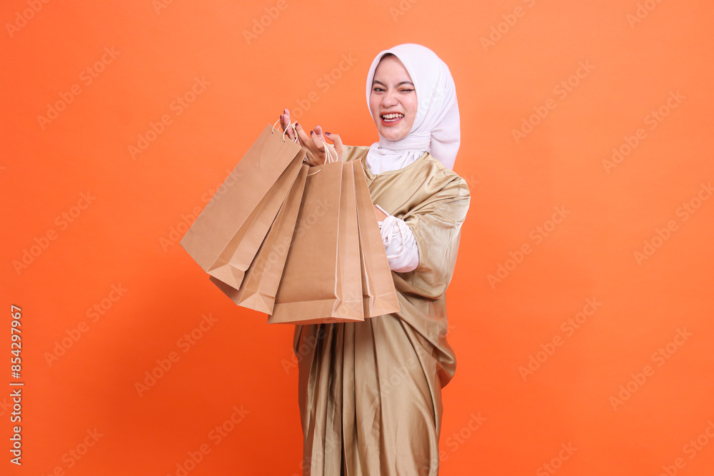 Cheerful mature Asian girl winks with both hands carrying a brown paper bag containing groceries in front of her wearing a kaftan Muslim dress with a hijab. Fashion concept, transaction, promotion