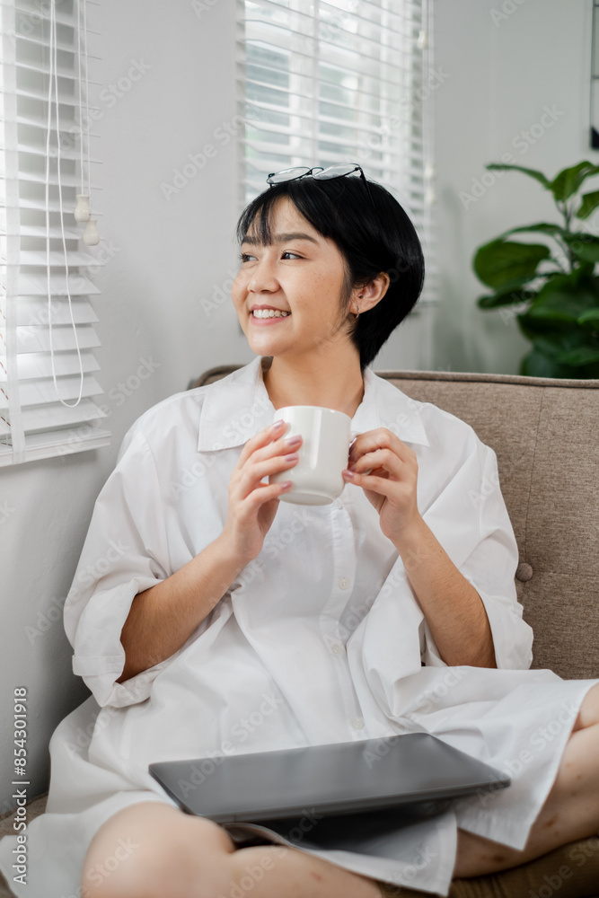 Young woman in white shirt holding coffee cup, sitting on a couch with a laptop, smiling and enjoying a peaceful morning at home. Young woman in white shirt holding coffee cup, sitting on a couch with a laptop, smiling and enjoying a peaceful morning at home.