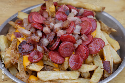 Fries. Closeup view of fried potatoes with sliced sausages and crispy bacon, in a metal dish with a wooden background.