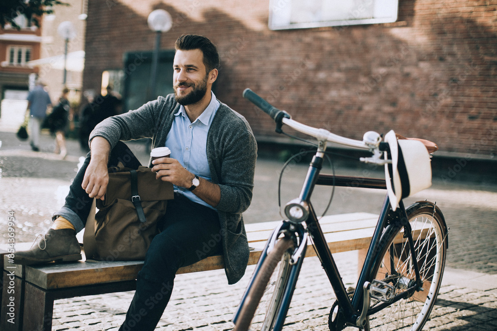 Obraz premium Relaxed stylish man sitting on bench with coffee next to bicycle in urban setting