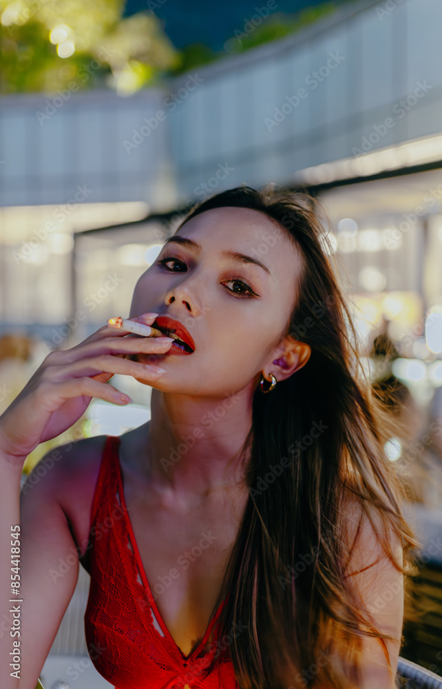 Fototapeta premium Sexy brunette asian woman tipsy in red dress blowing cigarette smoke on blurred cafe background at night.