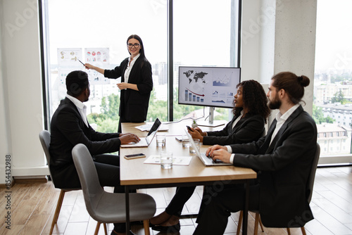 Business woman giving presentation to multiracial colleagues standing near graphs and charts on panoramic window. Professional team discussing growth statistics, global trends, and market analysis.
