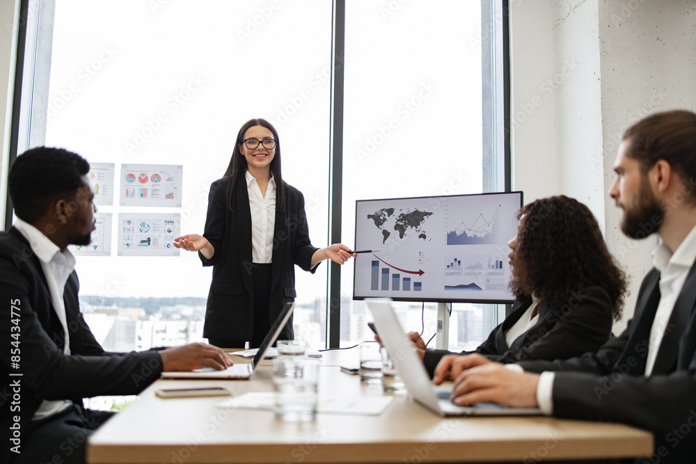 Business woman gives report to colleagues standing near graphs on monitor, screen, display, TV. Office setting with city view in background. Professional attire, teamwork, and corporate environment. Business woman gives report to colleagues standing near graphs on monitor, screen, display, TV. Office setting with city view in background. Professional attire, teamwork, and corporate environment.