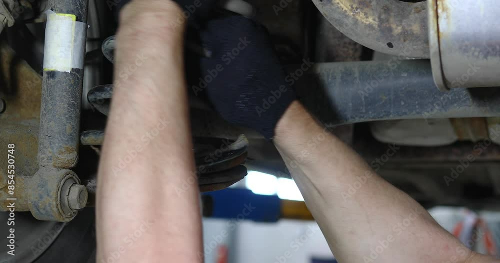 An auto mechanic removes a broken rear suspension spring of a passenger car for replacement, the workflow of servicing a vehicle at a technical inspection station An auto mechanic removes a broken rear suspension spring of a passenger car for replacement, the workflow of servicing a vehicle at a technical inspection station