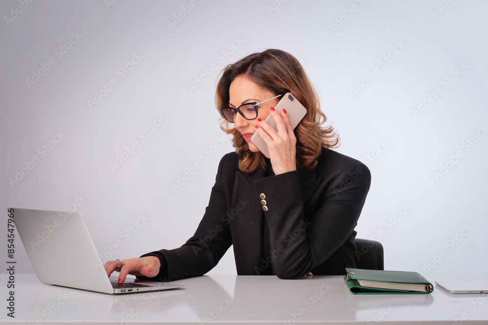 Mid aged businesswoman sitting at desk and using laptop and smartphone against isolated background Mid aged businesswoman sitting at desk and using laptop and smartphone against isolated background