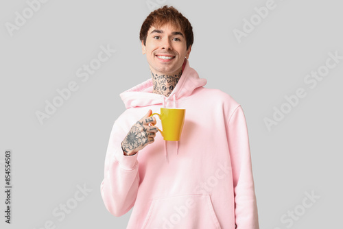 Happy handsome young tattooed man with cup of tea on grey background