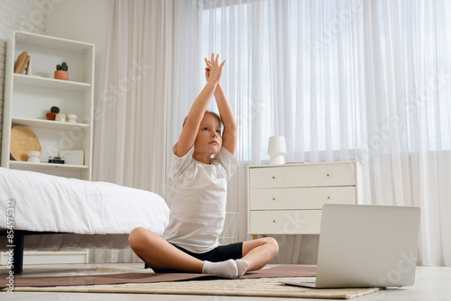 Little girl with mat and laptop doing stretching in bedroom