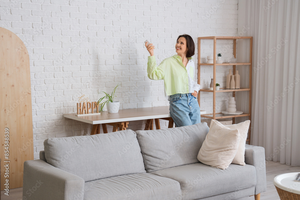 Smiling young woman with air conditioner remote control standing near sofa in living room Smiling young woman with air conditioner remote control standing near sofa in living room