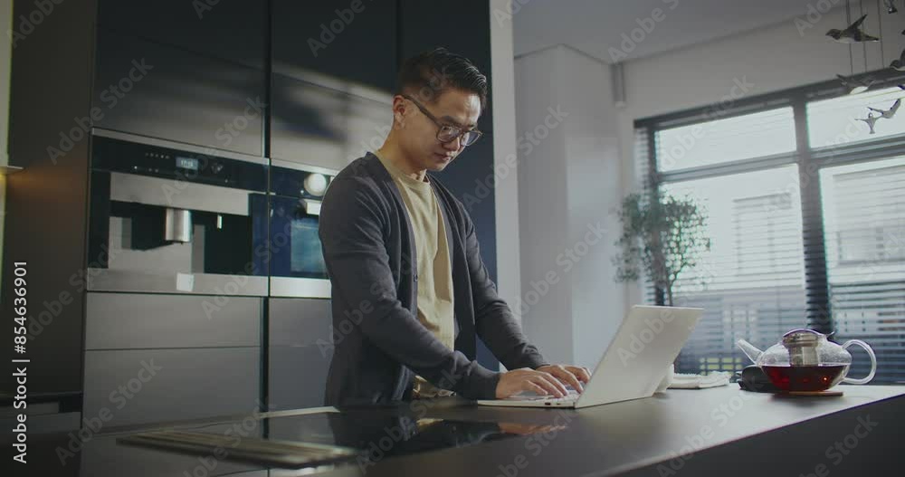 Handsome Asian man using laptop during his work. Working remotely with computer. Taking sip of hot drink. Feeling fresh during busy morning. Typing information on keyboard. Pressing keys. Handsome Asian man using laptop during his work. Working remotely with computer. Taking sip of hot drink. Feeling fresh during busy morning. Typing information on keyboard. Pressing keys.