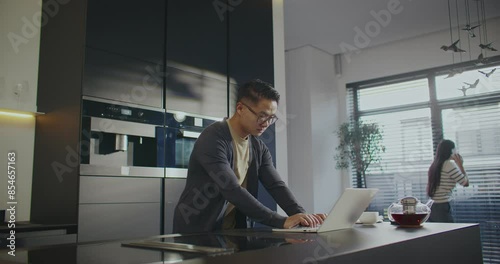 Hardworking freelancer using portative laptop for online work. Woman looking outdoor through window and talking with someone in background. Chinese couple with technology gadgets working from home. photo