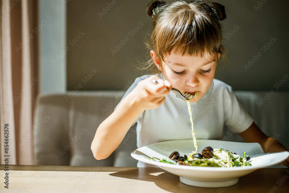 A cute little girl seated at a table, eating vegetarian pasta with green pesto and black olives A cute little girl seated at a table, eating vegetarian pasta with green pesto and black olives