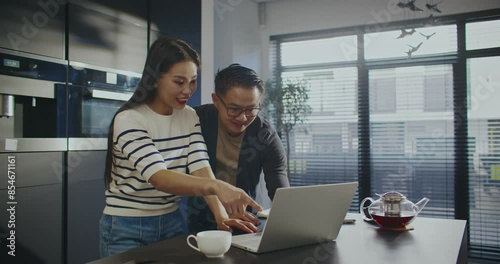 Lovely Asian couple standing in front of laptop. Woman opening lid of portable device. Waving their hands to relatives or friends. Video calling someone. Friendly gesture. Remote communication. photo