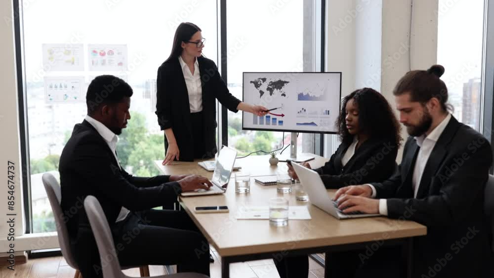 Business woman gives report to colleagues standing near graphs on monitor, screen, display, TV. Office setting with city view in background. Professional attire, teamwork, and corporate environment.