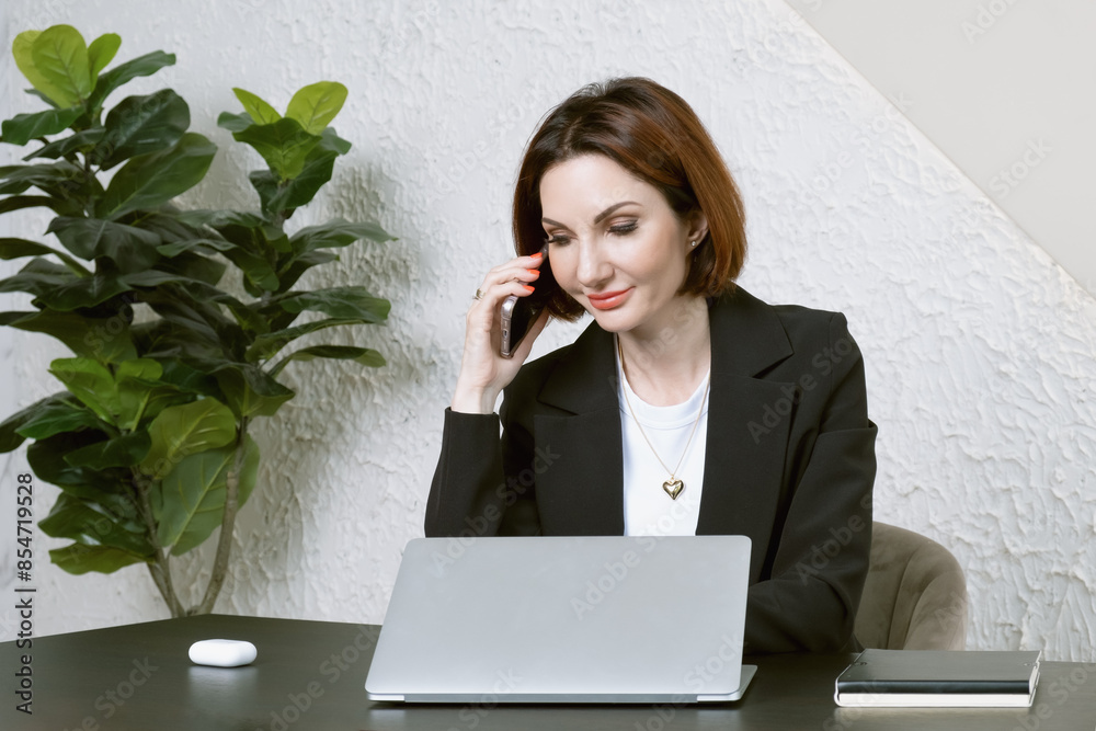 Portrait of businesswoman in the black jacket at desk in your office. Business portrait. Business woman is sitting in front of laptop screen and talking on cell phone Portrait of businesswoman in the black jacket at desk in your office. Business portrait. Business woman is sitting in front of laptop screen and talking on cell phone