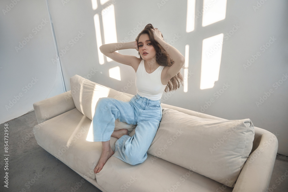 Young beautiful woman with long dark hair sits on white sofa, running her fingers through hair. Room is bright and sunny, with light streaming in through the window Young beautiful woman with long dark hair sits on white sofa, running her fingers through hair. Room is bright and sunny, with light streaming in through the window